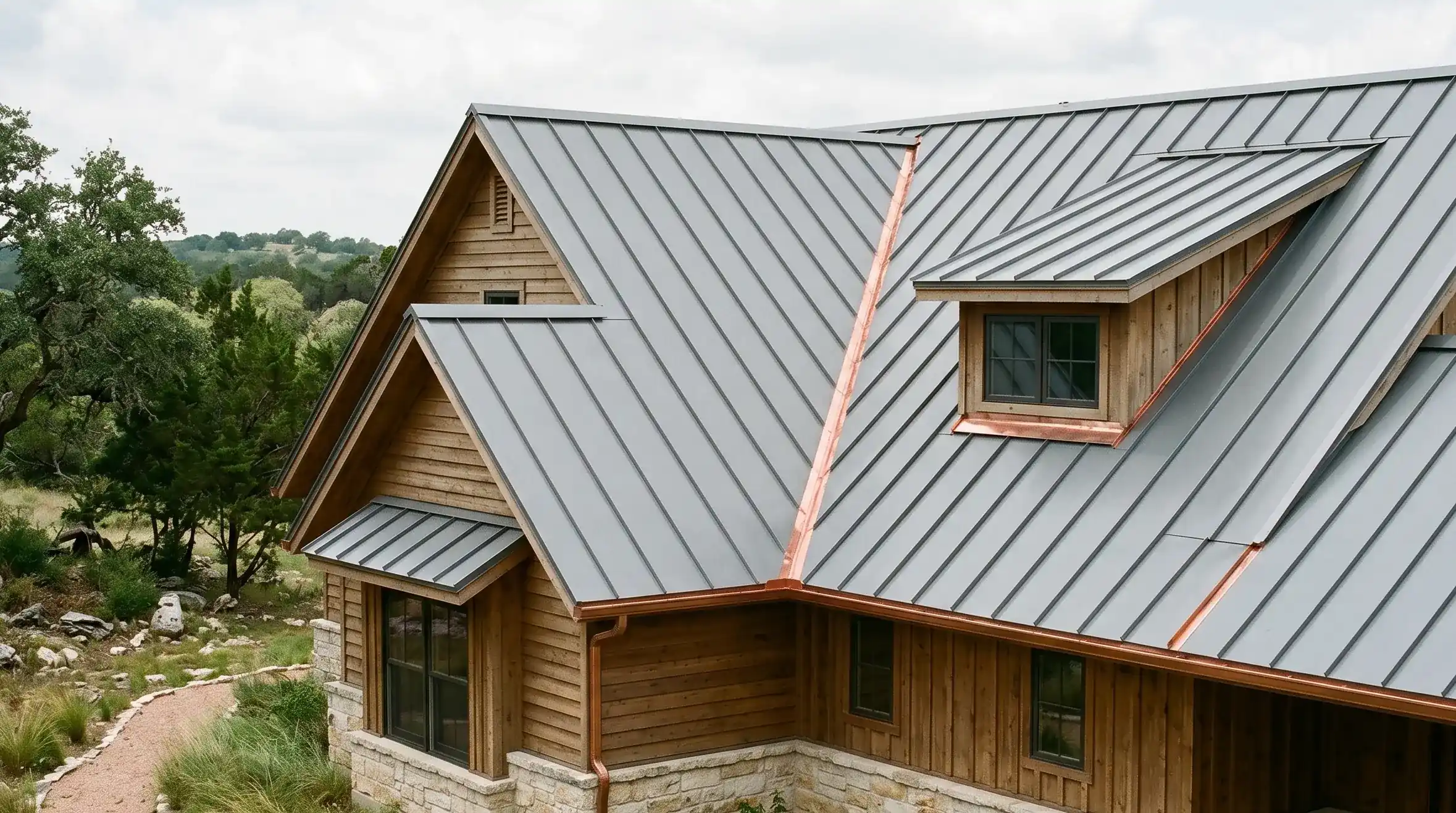 Wimberley custom home with new standing-seam metal roof at golden hour. Complex roofline with valleys and dormers visible; copper valley flashing catching the light.